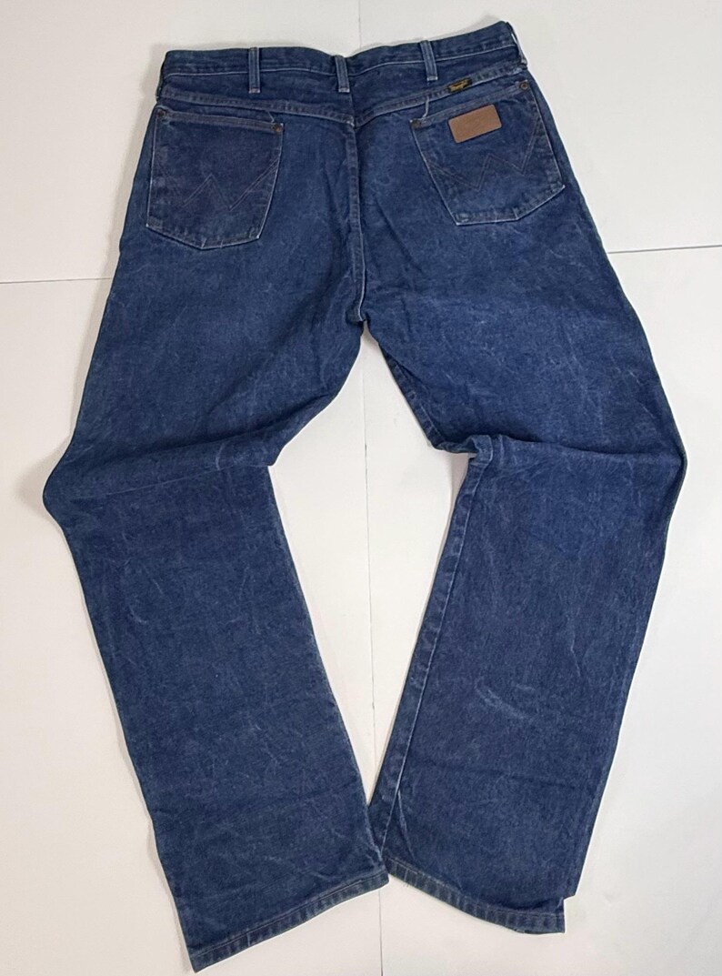 May include: Dark blue denim jeans with a classic five-pocket design. The jeans have a straight leg cut and a brown leather patch on the back pocket. The Wrangler logo is visible on the patch. The jeans are laid flat on a white surface.