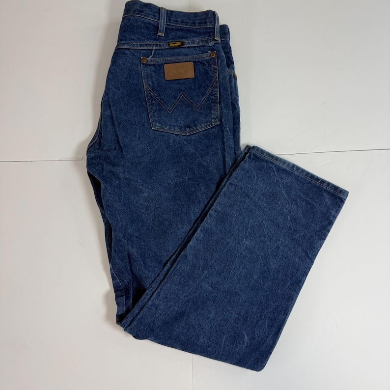 May include: Blue denim Wrangler jeans. The jeans have a classic five-pocket design, a brown leather patch on the back, and the Wrangler logo. The jeans are folded on a white surface.
