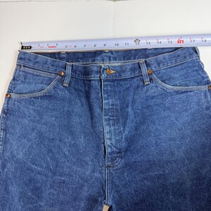 May include: Close-up of blue denim jeans with a button closure and pockets. The jeans are measured with a tape measure, showing a waist size of approximately 40.6 cm.