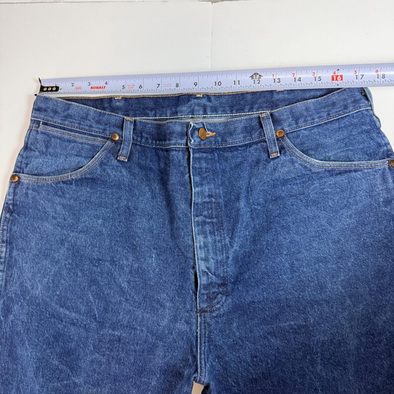 May include: Close-up of blue denim jeans with a button closure and pockets. The jeans are measured with a tape measure, showing a waist size of approximately 40.6 cm.