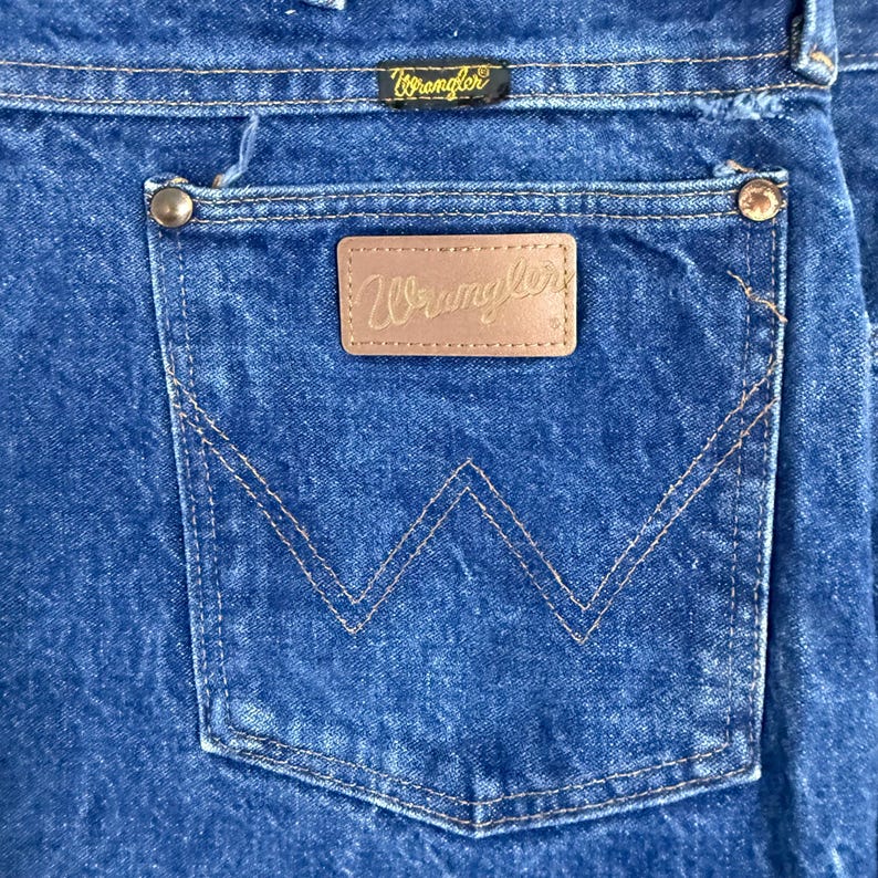 May include: Close-up of the back pocket of blue denim jeans. The pocket features a brown leather patch with the "Wrangler" logo and a stitched "W" design. The "Wrangler" brand name is also visible above the pocket.