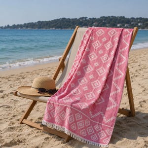 May include: A pink and white patterned beach towel with a geometric design and fringe edges draped over a wooden beach chair, accompanied by a tan straw hat with black sunglasses resting on the brim, set against a sandy beach background.