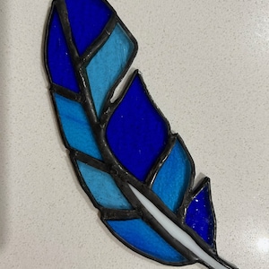 May include: A stained glass feather with a blue and white design. The feather is made of glass pieces that are joined together with black lead. The feather has a white line down the center.