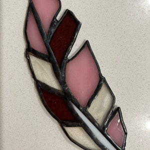 May include: A stained glass feather ornament with pink, white, and burgundy glass pieces. The feather is outlined in black metal and has a small loop at the bottom for hanging.