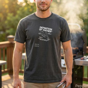 May include: A charcoal gray short-sleeve t-shirt featuring white text that reads DEFINITION OF FATHER with a diagram of a hammer, wrench, and coffee mug. Below the diagram, the text reads PROVIDER, PROTECTOR, GUIDE, ESSENTIAL FUNCTIONS.