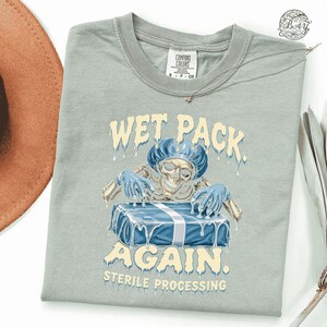 May include: A light gray crew neck t-shirt featuring a graphic of a skeleton wearing a blue surgical cap and gloves, reaching for a blue wrapped package. The text reads WET PACK AGAIN STERILE PROCESSING in a dripping, cream-colored font.