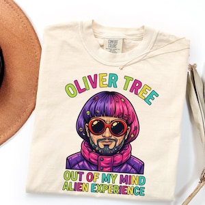 May include: A cream-colored t-shirt featuring a graphic of Oliver Tree with a purple and pink bowl cut, wearing red-rimmed sunglasses and a purple jacket. The text reads OLIVER TREE above and OUT OF MY MIND ALIEN EXPERIENCE below in colorful letters.