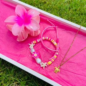 Könnte beinhalten: Eine rosa und weiße Blumenhaarklammer, ein rosa und gelbes Perlenarmband mit weißen Perlenakzenten und eine goldene Schildkrötenkette auf rosa Untergrund.