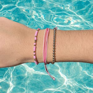 Könnte beinhalten: Drei Armbänder an einem Handgelenk. Eines ist ein rosafarbenes Perlenarmband, eines ist ein weiß-rosa Schnurarmband und eines ist ein silbernes Perlenarmband.