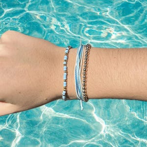 Könnte beinhalten: Drei Armbänder an einem Handgelenk. Die Armbänder sind aus blauem und weißem Band, blauen und goldenen Perlen und goldenen Perlen.