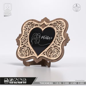 Può includere: Cornice in legno per la festa della mamma con un cuore al centro. La cornice presenta un design floreale tagliato al laser e un cuore nero con un'illustrazione di madre e figlio e la scritta "Happy Mother's Day". La cornice è spessa 3 mm.