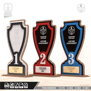 May include: Three wooden trophies, numbered 1, 2, and 3, with a shield-shaped design. Each trophy has a different color scheme: black and white, red, and blue. The text "PRESENTED FOR YOUR LIFETIME ACHIEVEMENT" is displayed on each trophy. The base includes space for a name and the year 2026.