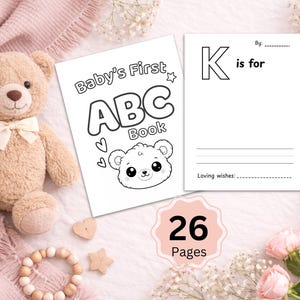 Może przedstawiać: Miś w kolorze beżowym z kremową kokardą leży obok książeczki do kolorowania ABC dla dziecka. Książka zawiera ilustrację misia i napis "Baby's First ABC Book". Strona głosi "K is for". Obecne są również drewniane serce, gwiazda i bransoletka.