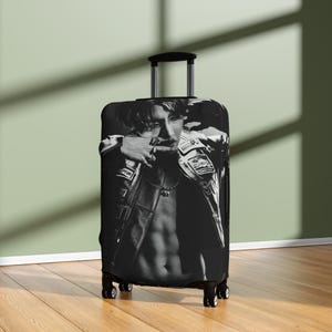 以下が含まれることがあります： 黒と白のスーツケースカバー。革のジャケットを着た人物が描かれています。画像は、ジャケットとネックレスが見える、人物の上半身のクローズアップです。スーツケースには格納式のハンドルと車輪が付いています。