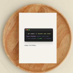 May include: A white greeting card with the text "happy birthday." The card features a simulated code snippet with the text "git commit -m 'another year older'" and other lines of code. The card is on a wooden plate.