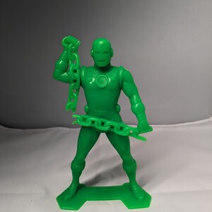 May include: Green plastic action figure of a superhero holding a chain in one hand. The figure is standing on a green plastic base.