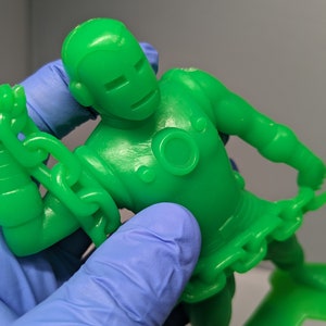 May include: A green plastic action figure of a superhero wearing a chain around their waist and arms. The figure is posed with one arm raised and the other arm bent at the elbow.