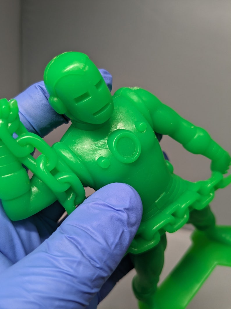 May include: A green plastic action figure of a superhero wearing a chain around their waist and arms. The figure is posed with one arm raised and the other arm bent at the elbow.