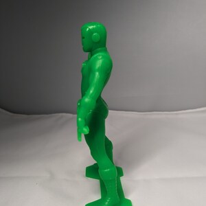 May include: A green plastic action figure of a superhero standing on a base. The figure has a helmet and is wearing a suit with a belt.