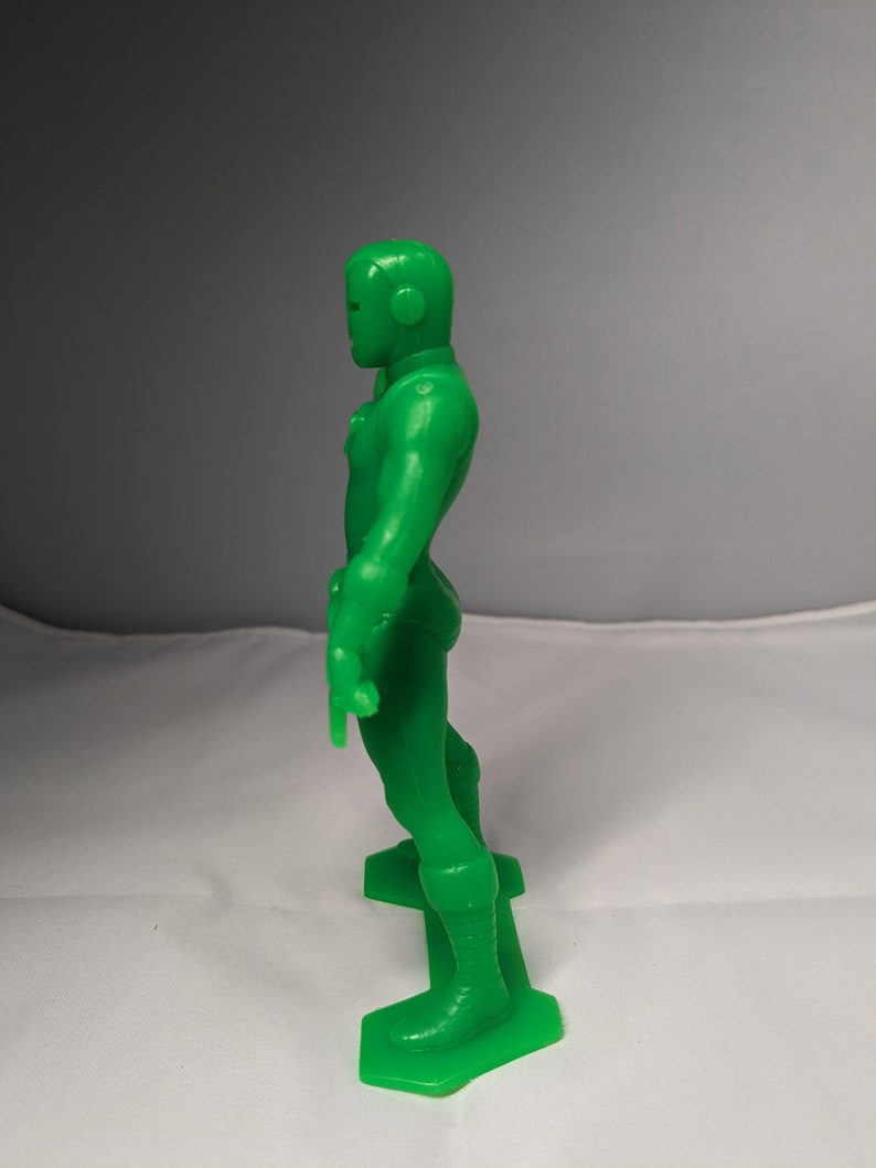 May include: A green plastic action figure of a superhero standing on a base. The figure has a helmet and is wearing a suit with a belt.