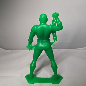 May include: A green plastic action figure of a superhero holding a chain in one hand. The figure is standing on a green plastic base.