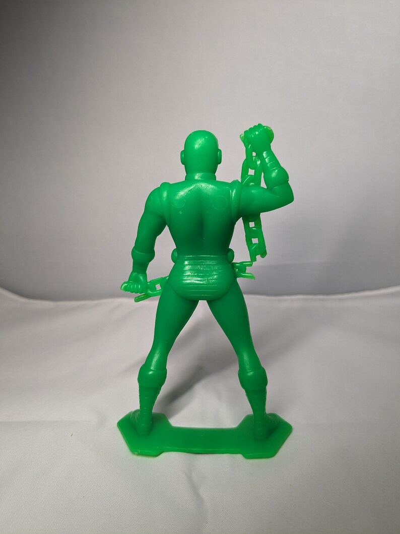 May include: A green plastic action figure of a superhero holding a chain in one hand. The figure is standing on a green plastic base.