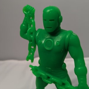 May include: Green plastic action figure of a superhero holding a chain in each hand. The figure has a metallic look and a helmet with a face mask.