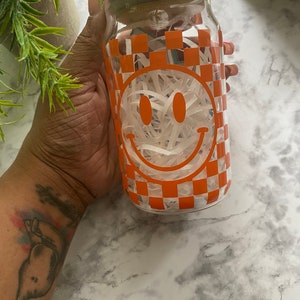 K&ouml;nnte beinhalten: Ein klares Glasgef&auml;&szlig; mit einem Holzdeckel und einem karierten Muster mit einem Smiley-Gesicht. Das Gef&auml;&szlig; ist mit wei&szlig;em zerkleinertem Papier gef&uuml;llt.