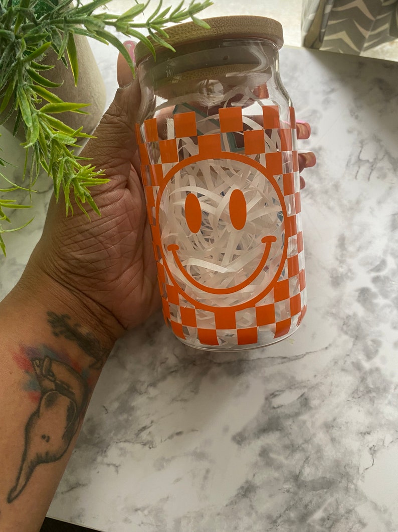 K&ouml;nnte beinhalten: Ein klares Glasgef&auml;&szlig; mit einem Holzdeckel und einem karierten Muster mit einem Smiley-Gesicht. Das Gef&auml;&szlig; ist mit wei&szlig;em zerkleinertem Papier gef&uuml;llt.