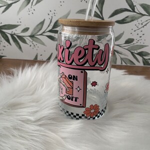 May include: A glass tumbler with a bamboo lid and a straw. The tumbler has a pink and black design with the word "anxiety" and a toggle switch that says "on" and "off".