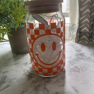 K&ouml;nnte beinhalten: Ein klares Glasgef&auml;&szlig; mit einem Holzdeckel, gef&uuml;llt mit wei&szlig;en Papierschnitzeln. Das Gef&auml;&szlig; hat ein kariertes Muster in Orange und Wei&szlig; mit einem wei&szlig;en Smiley-Gesicht.