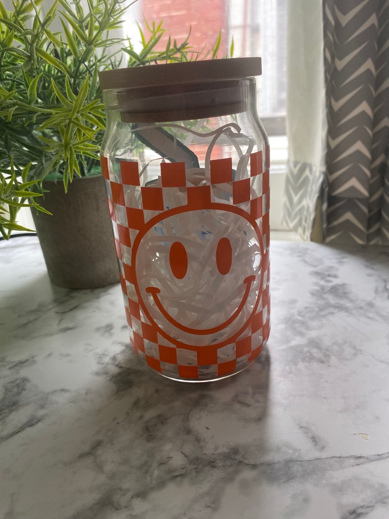 K&ouml;nnte beinhalten: Ein klares Glasgef&auml;&szlig; mit einem Holzdeckel, gef&uuml;llt mit wei&szlig;en Papierschnitzeln. Das Gef&auml;&szlig; hat ein kariertes Muster in Orange und Wei&szlig; mit einem wei&szlig;en Smiley-Gesicht.