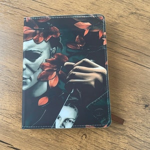 May include: A black leather notebook featuring a graphic print of a horror movie character holding a knife, with red leaves obscuring part of their face.