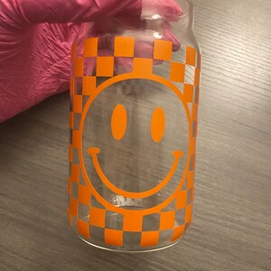 K&ouml;nnte beinhalten: Ein klares Glasgef&auml;&szlig; mit einem karierten Muster und einem gro&szlig;en orangefarbenen Smiley-Gesicht.