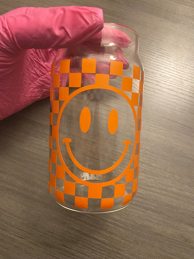 K&ouml;nnte beinhalten: Ein klares Glasgef&auml;&szlig; mit einem karierten Muster und einem gro&szlig;en orangefarbenen Smiley-Gesicht.