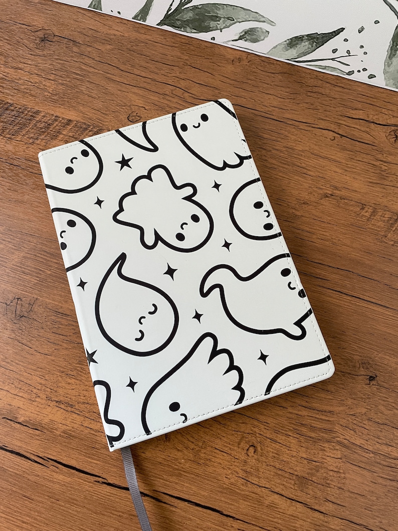 May include: A white notebook with a black and white ghost pattern. The notebook has a grey ribbon bookmark.