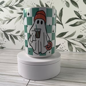 Puede incluir: Taza de cerámica blanca con un patrón de cuadros verde y blanco. Un fantasma de dibujos animados con un sombrero naranja sostiene una taza de café y una pequeña calabaza. La taza tiene un borde y un asa blancos.