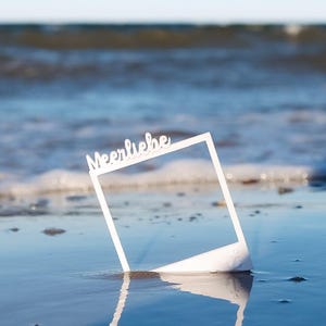 Könnte beinhalten: Ein weißer, quadratischer Fotorahmen-Ausschnitt mit dem Wort Meerliebe in Schreibschrift am oberen Rand. Der Rahmen steht aufrecht auf einem nassen Sandstrand, wobei die Meereswellen und ein klarer blauer Himmel im Hintergrund zu sehen sind.