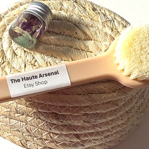 May include: A close-up of a light pink facial brush with white bristles. The brush is lying on a woven straw basket. A small glass jar with a silver lid and purple flowers inside is also in the basket. The jar and brush are labeled "The Haute Arsenal Etsy Shop".