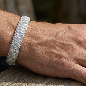 以下が含まれることがあります： 小さな輝く石が多数セットされた、パヴェデザインのシルバーブレスレット。 ブレスレットは幅が広く、手首に巻き付いています。 画像は、人の腕にブレスレットを示しています。