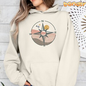 May include: A cream-colored hoodie with a compass design featuring a black compass rose, a pink and brown mountain range, and a yellow sun. The compass points are labeled N, S, E, and W.