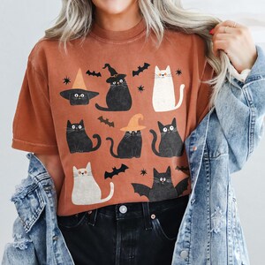 May include: A rust-colored t-shirt with a graphic print of black cats wearing witch hats and a white cat. The cats are surrounded by bats and stars.