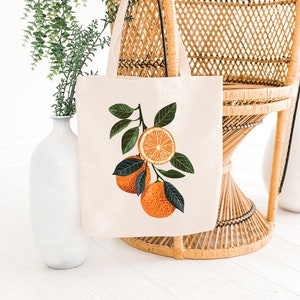 May include: A white canvas tote bag with a design of orange slices and green leaves. The bag is sitting on a wicker chair.