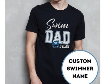 May include: Black t-shirt with white text that reads "Swim Dad" and a blue swimmer graphic with the name "Dylan" underneath. The text "Custom Swimmer Name" is in a circle on the right side of the shirt.