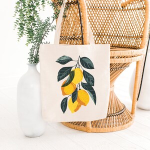 May include: A white tote bag with a lemon branch design. The bag has a yellow, green, and white color scheme. The bag is sitting on a wicker chair.