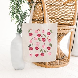 May include: A white canvas tote bag with a repeating pattern of pink and red cherries. The bag is hanging on a wicker chair.