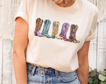 May include: A white t-shirt with a graphic of five colorful cowboy boots. The boots are arranged in a row, with the toes pointing to the right. The boots are all different colors, including brown, blue, yellow, purple, and pink.