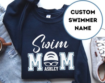 May include: Navy blue sweatshirt with the text "Swim Mom" in white with a swimmer graphic and the name "Ashley" underneath. The sweatshirt is on a white background with a pair of blue jeans and a cup of coffee.
