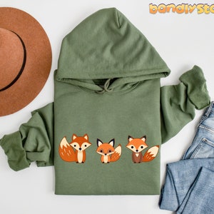 May include: A green hooded sweatshirt with a graphic of three orange foxes with white accents. The foxes are facing forward with their tails curled behind them.