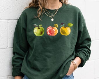 May include: A forest green long-sleeve sweatshirt featuring a graphic print of three apples in green, red, and yellow. The sweatshirt has a crew neckline and is paired with blue denim jeans. The text bandlystore is visible in the top left corner.